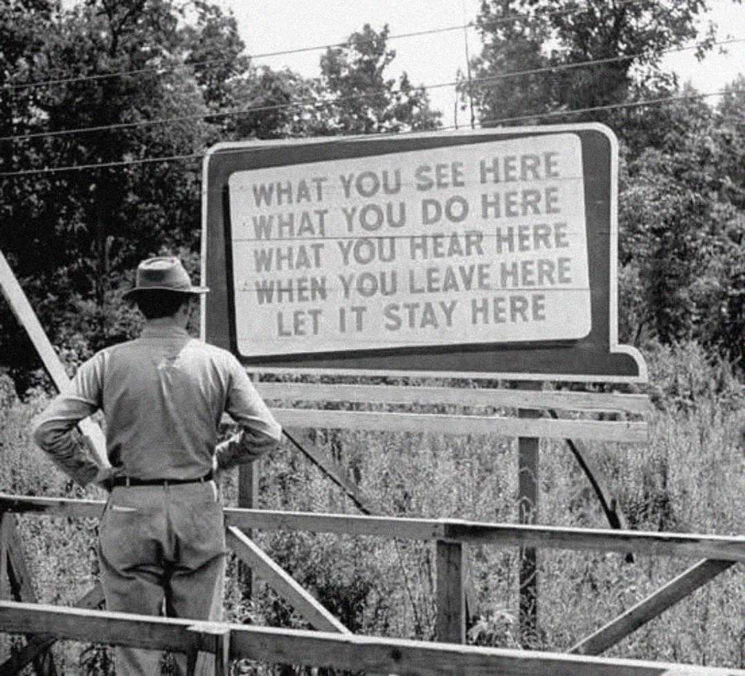 A man in a hat and uniform stands in front of a large sign that reads, “WHAT YOU SEE HERE, WHAT YOU DO HERE, WHAT YOU HEAR HERE, WHEN YOU LEAVE HERE, LET IT STAY HERE." Trees and bushes are in the background.