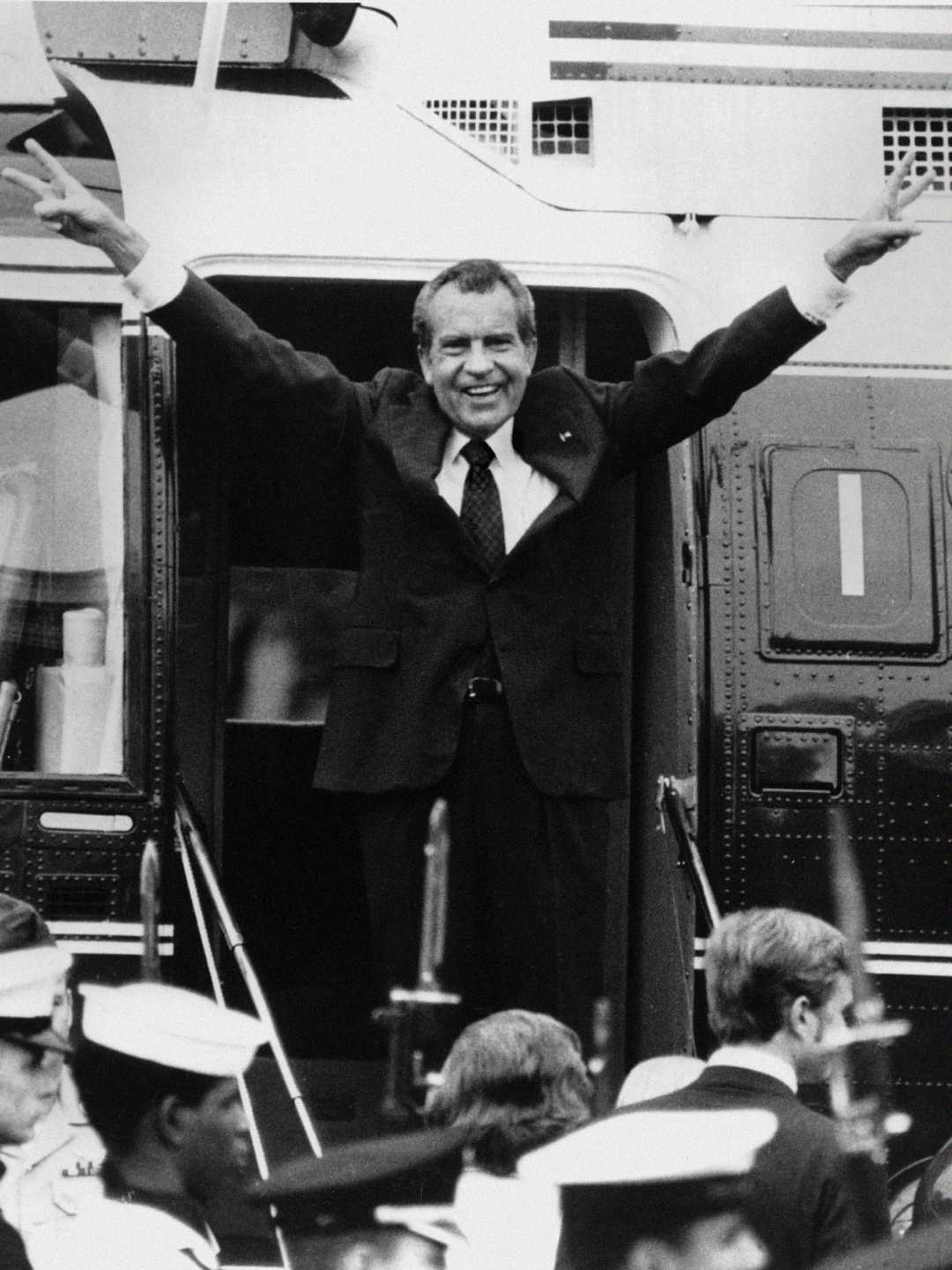 A man in a suit stands at the door of a helicopter, smiling and raising both arms with fingers forming V-signs. People are gathered below, some in military uniforms, watching him.