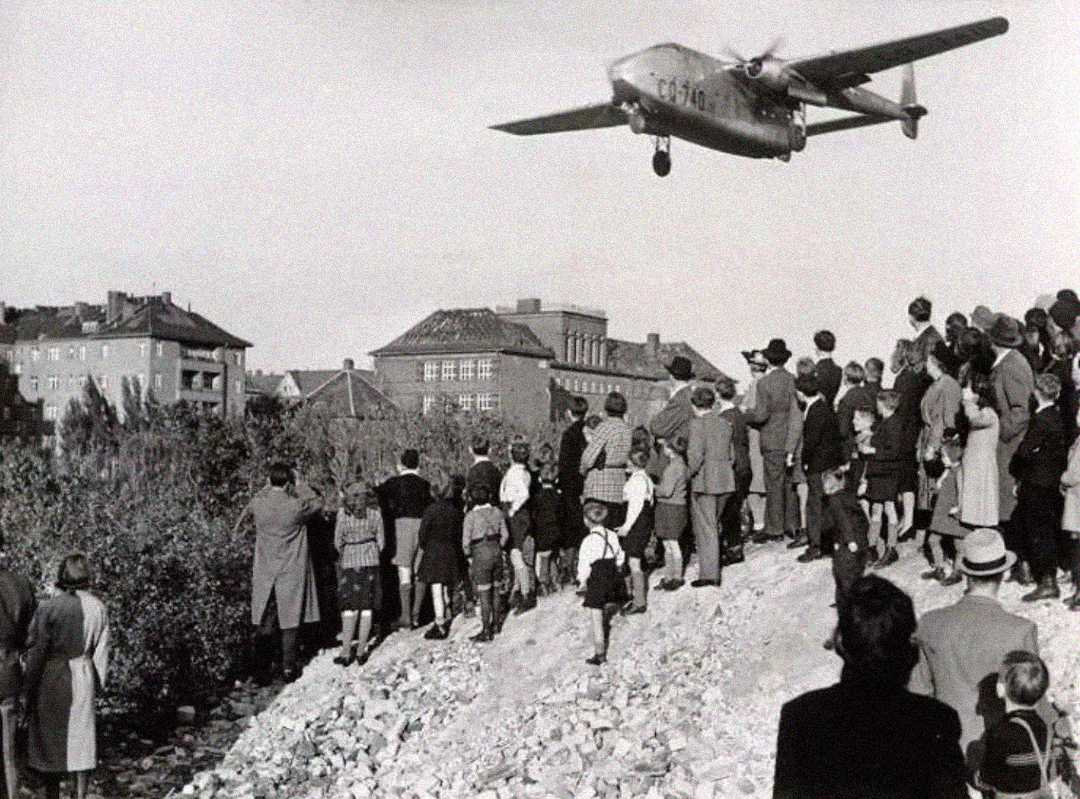 A black-and-white photo shows a crowd of people watching a low-flying airplane pass over a cityscape. The group stands on a rocky mound, while buildings and trees are visible in the background.