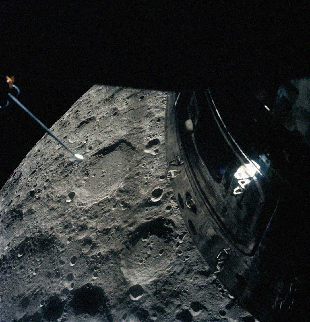 A spacecraft orbits above the cratered surface of the Moon, with part of the spacecraft and a long instrument or antenna visible in the foreground. The lunar surface is covered in numerous craters.