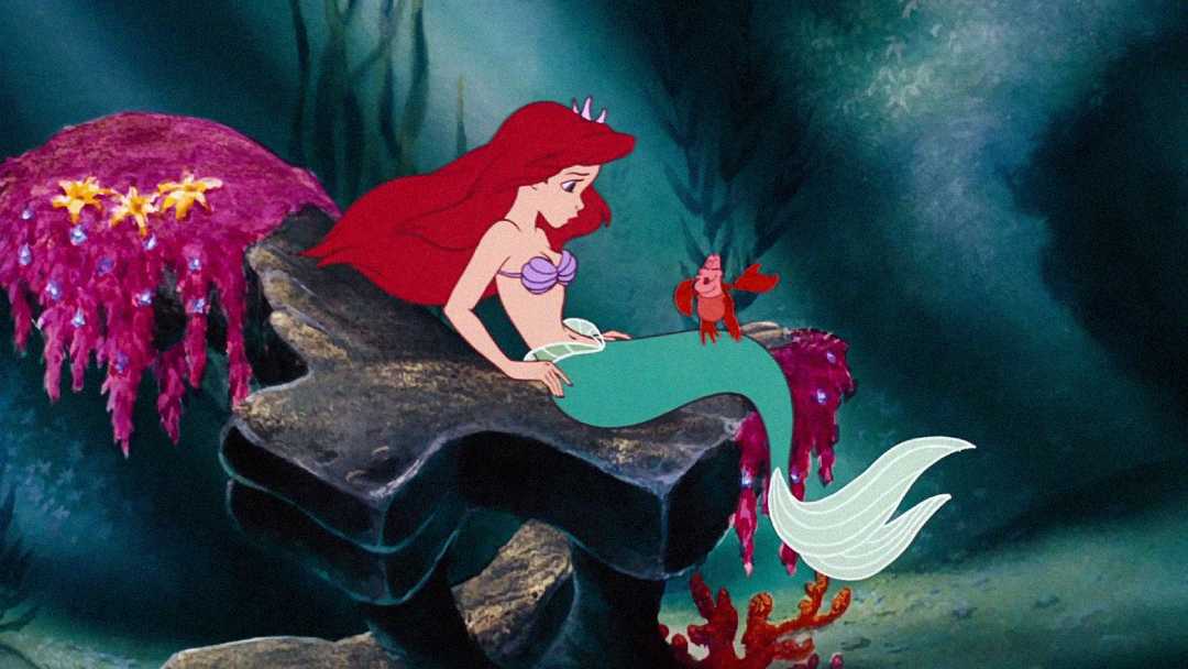 Animated mermaid with long red hair sits on a rock underwater, talking to a red crab. Colorful sea plants and coral surround them in the scene.