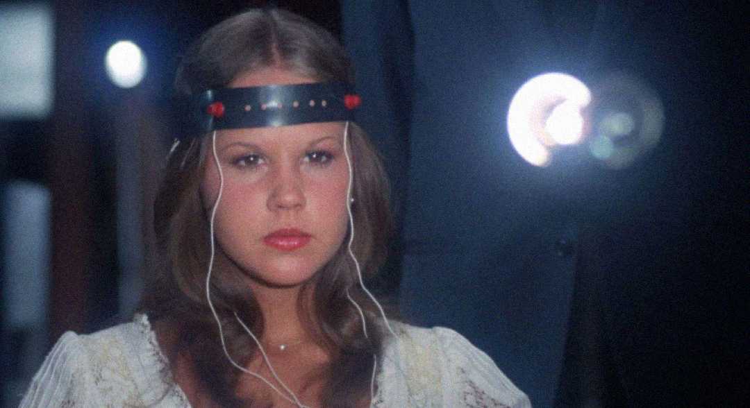 A woman with long brown hair wears a metallic headband with wires attached, looking serious. She sits in a dimly lit room with a circular light glowing in the background.