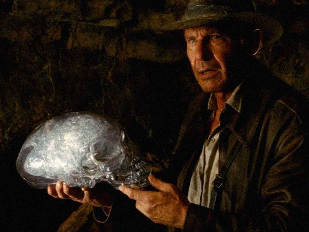 A man in a brown leather jacket and hat holds a large, transparent crystal skull with both hands, standing in a dimly lit, rocky environment.