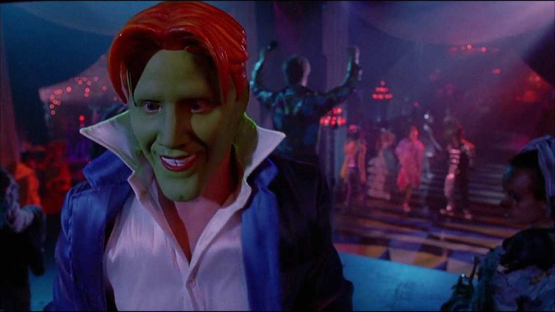 A person with bright green skin, red hair, and exaggerated facial features wears a blue jacket and white shirt, grinning at the camera in a dimly lit party scene with people dancing in the background.