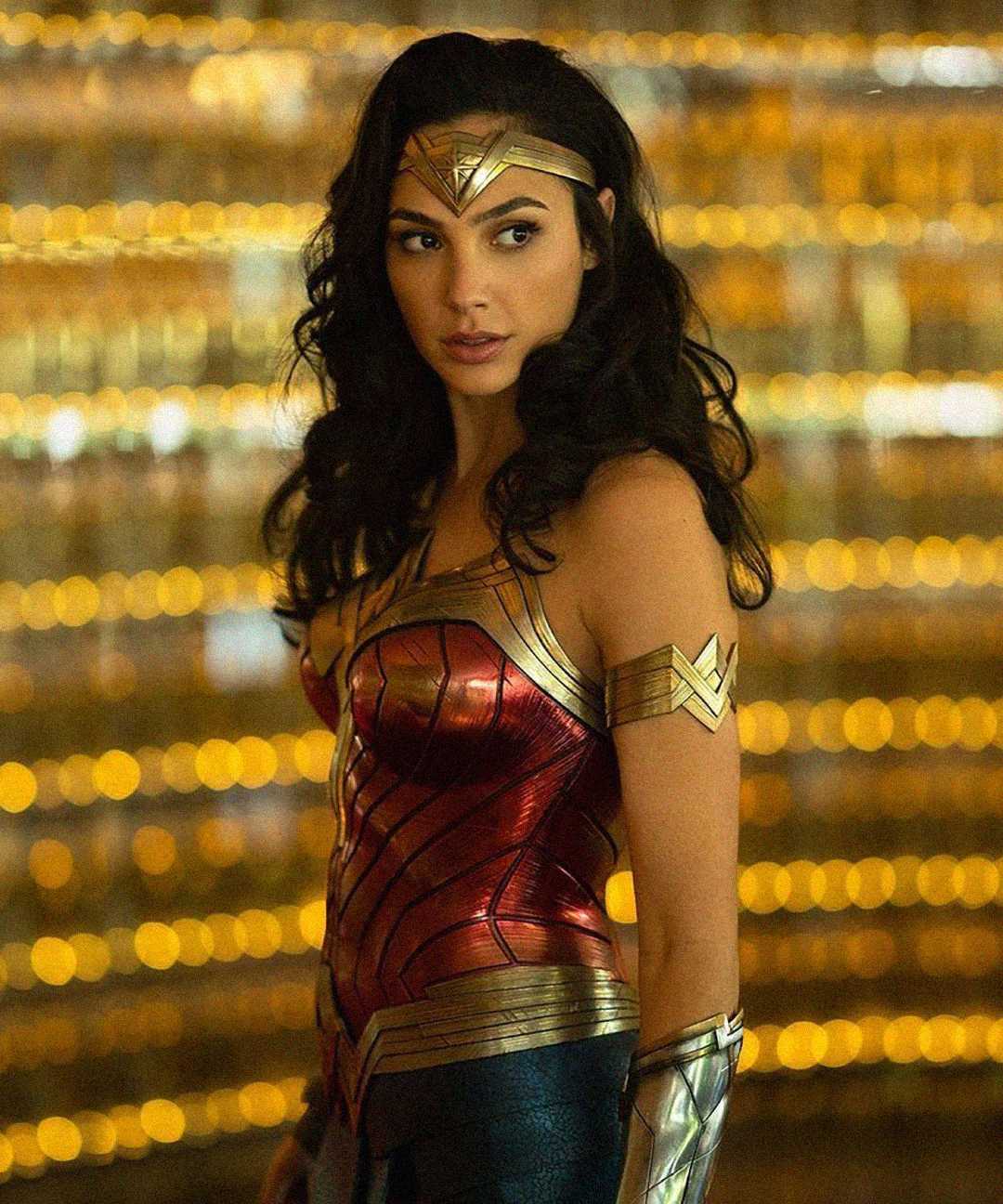 A woman in a red and gold superhero costume with a tiara stands against a background of golden lights, looking to the side with a serious expression.
