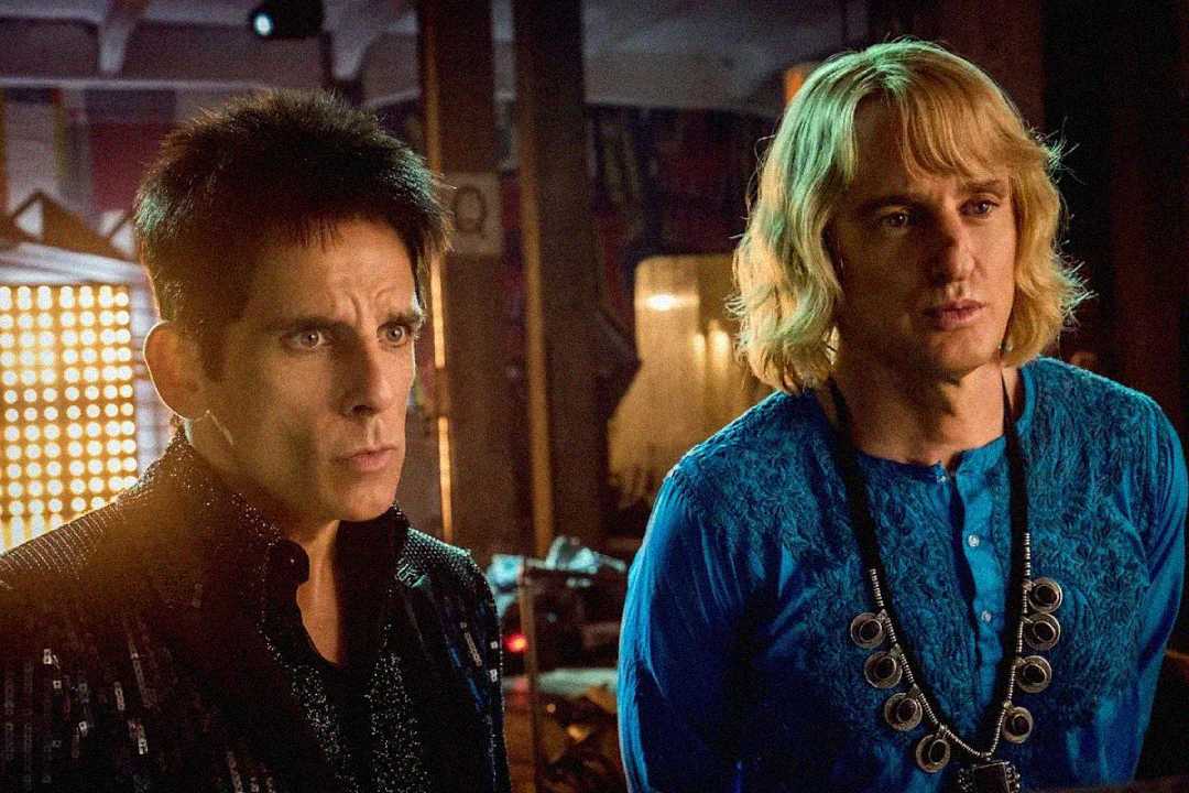 Two men stand side by side indoors; one has short dark hair and wears a black outfit, while the other has long blond hair and wears a bright blue shirt with a large necklace. Both have serious expressions.