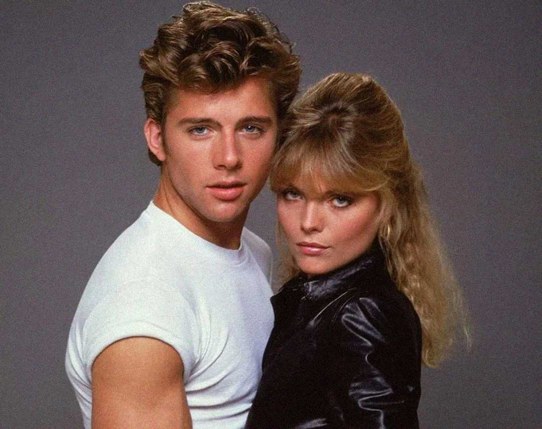 A young man in a white T-shirt stands closely beside a young woman with long blonde hair, wearing a black leather jacket. Both look directly at the camera with serious expressions against a plain gray background.