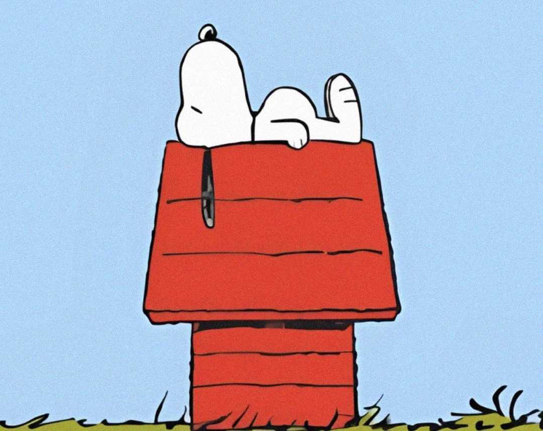 A cartoon dog is lying on its back, relaxing on the roof of a red doghouse, with grass below and a clear blue sky in the background.