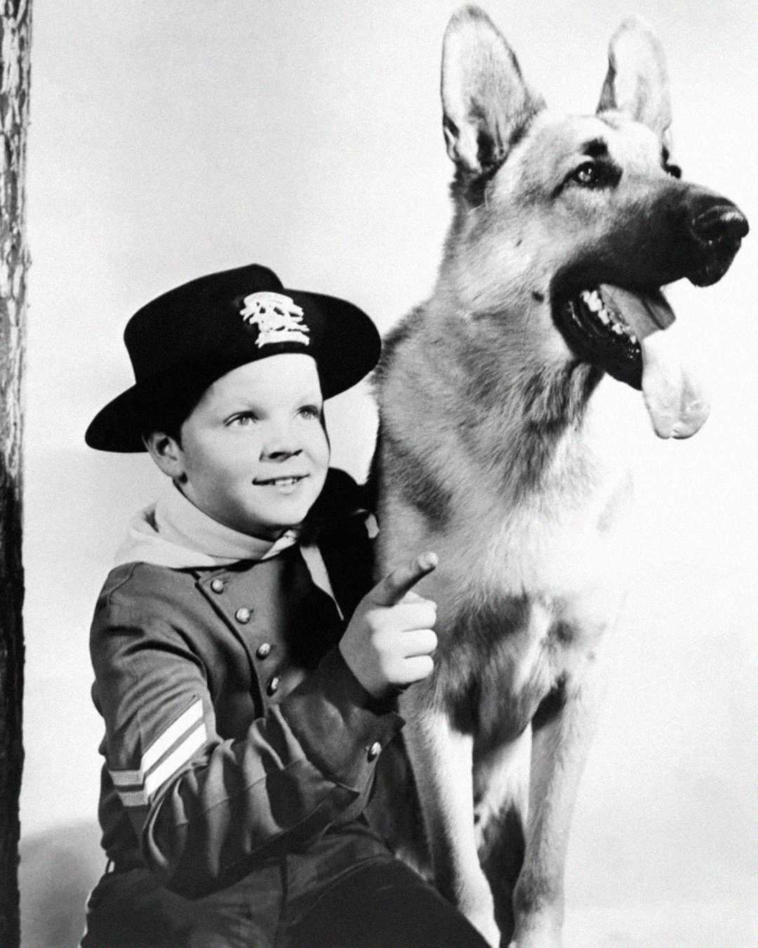 A young boy in a uniform and hat sits next to a large German Shepherd dog. The boy is smiling and pointing upwards, while the dog sits beside him with its tongue out. The image is in black and white.