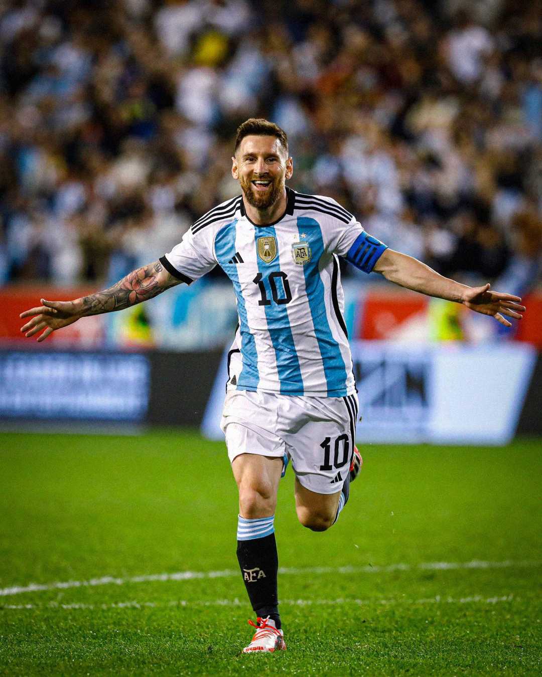 A soccer player in an Argentina jersey with number 10 celebrates on the field with arms outstretched, smiling, as a crowd of fans cheers in the blurred background.