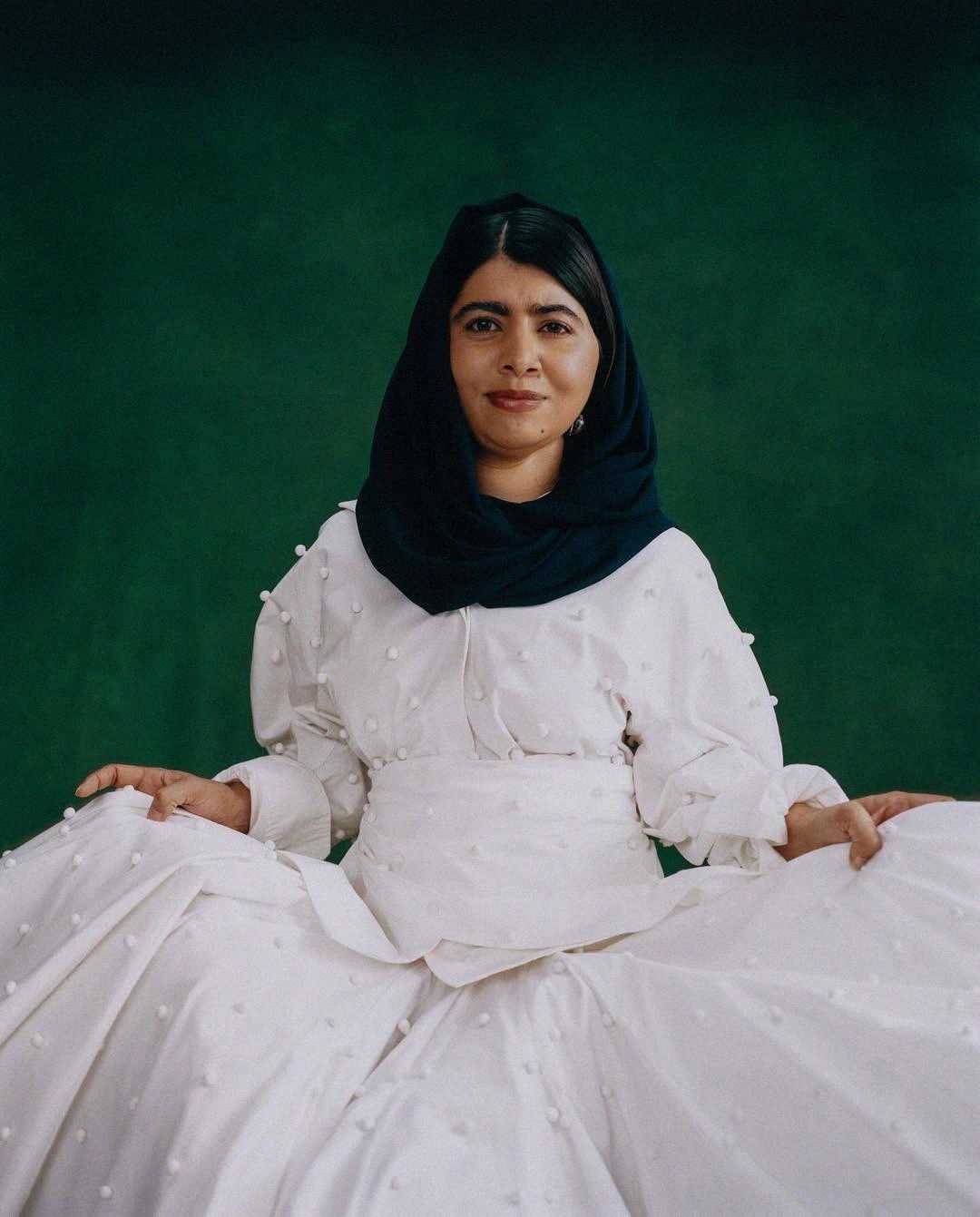 A woman wearing a dark headscarf and a long white dress with textured details sits against a dark green background, holding the sides of her dress and looking confidently at the camera.