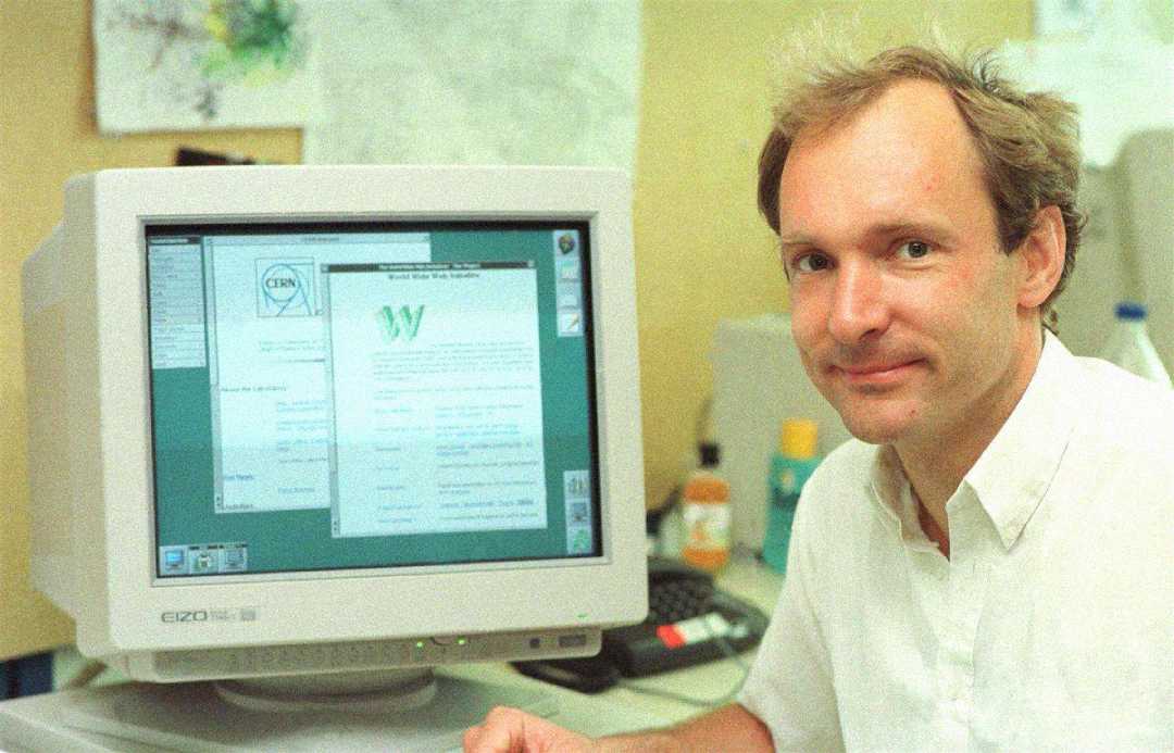 A man in a white shirt sits beside an old computer monitor displaying an early web browser interface, with papers and office supplies in the background.