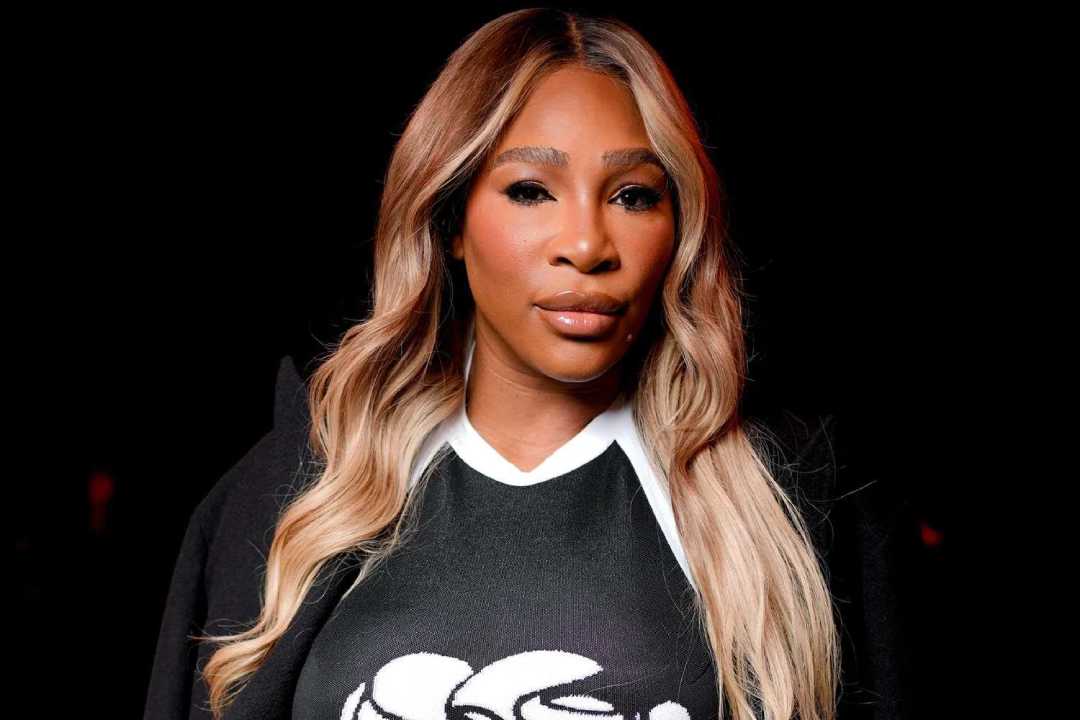 A woman with long, wavy blonde hair wearing a black and white top stands against a dark background, looking confidently at the camera.