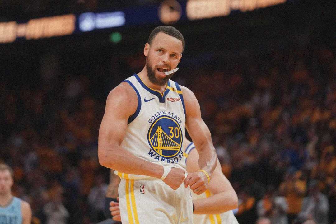 A basketball player in a Golden State Warriors jersey flexes his arms and celebrates on the court, with a mouthguard hanging from his mouth. The background shows a blurred crowd and fellow players.