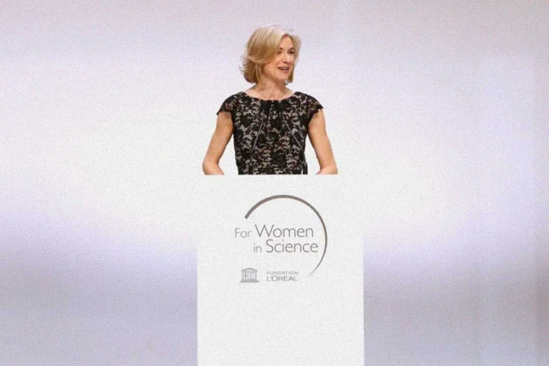 A woman in a black lace dress stands at a podium with the logo "For Women in Science" and "Fondation L'Oréal" printed on it, speaking at a formal event.