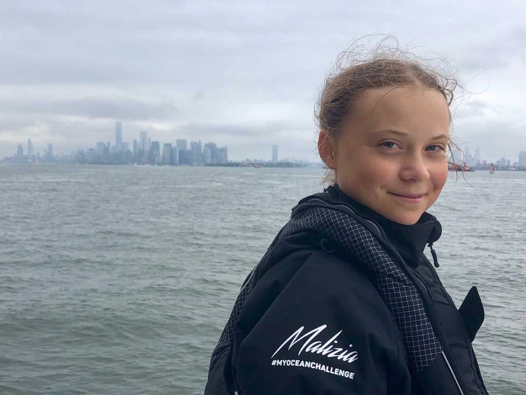 A young person with light brown hair stands by the water, wearing a dark jacket that says "Malizia #MYOCEANCHALLENGE." A city skyline with tall buildings is visible in the background under a cloudy sky.