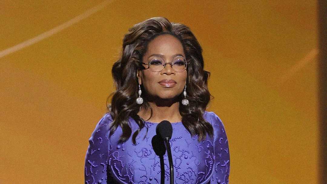 A woman in a purple embroidered dress and round glasses stands at a microphone against a warm, golden background, wearing dangling earrings and looking forward.