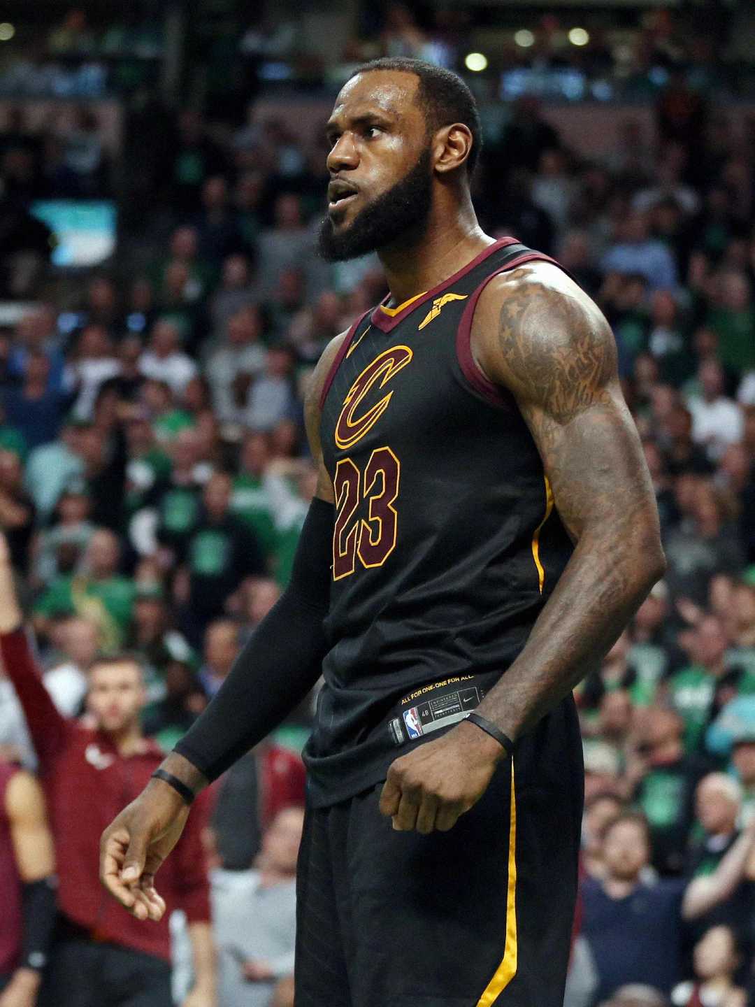 A basketball player in a Cleveland Cavaliers jersey with the number 23 stands on the court, looking focused. The arena background is filled with spectators.