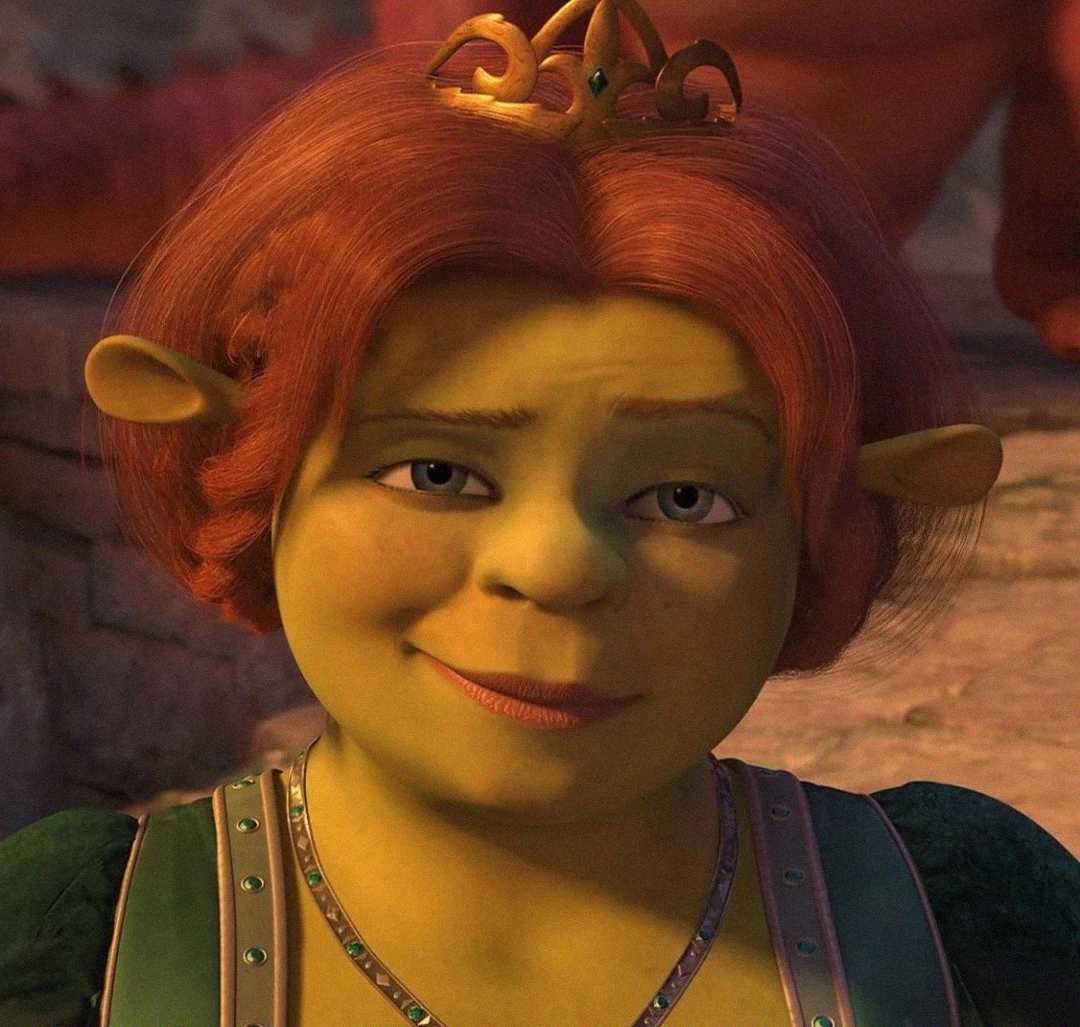 Princess Fiona, an ogre with green skin, red hair, and wearing a small gold crown and medieval-style dress, smiles softly while looking slightly off to the side. The background is softly blurred.