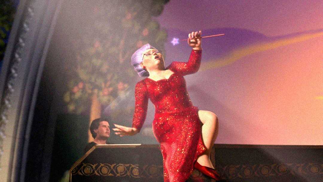A fairy godmother with purple hair and glasses, wearing a sparkling red dress, dances and sings joyfully on stage while holding a glowing wand. A man is seen in the background at a piano.