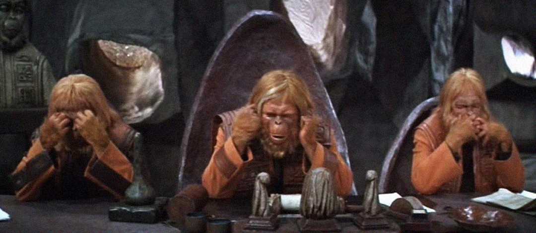 Three humanoid apes in robes mimic "see no evil, hear no evil, speak no evil" gestures while sitting at a stone table in a dimly lit, cave-like room.