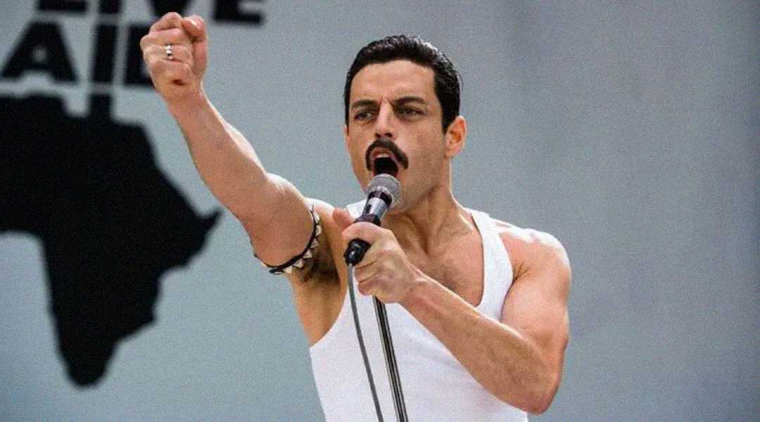 A man in a white tank top passionately sings into a microphone, raising his fist, with a map of Africa partially visible in the background.