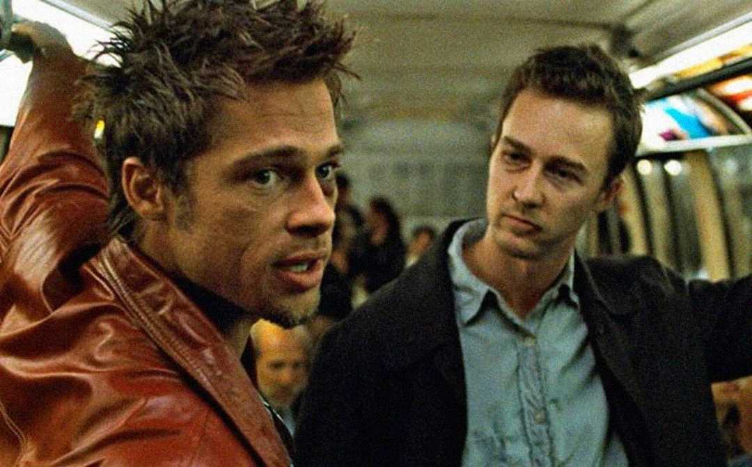 Two men stand in a crowded subway car. One wears a red leather jacket and looks over his shoulder, while the other, in a collared shirt, gazes seriously at him. The scene appears tense and dramatic.
