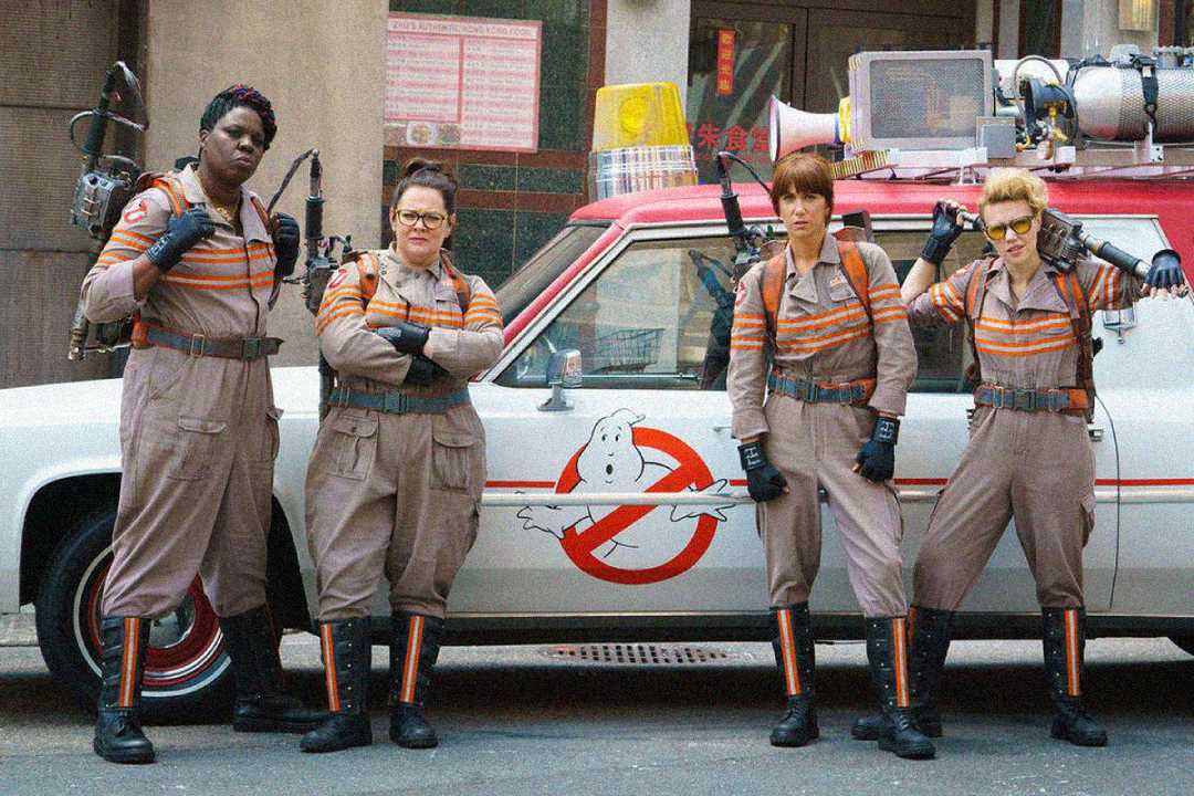 Four women in Ghostbusters uniforms stand confidently in front of the Ghostbusters car, with proton packs on their backs, posing for the camera on a city street.