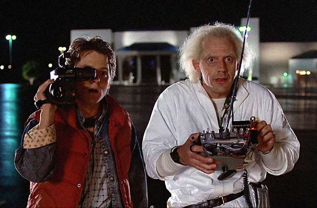 Two men stand outside at night; one holds a video camera, and the other, with wild white hair, wears a white jumpsuit and holds a remote control. Both look surprised, with a mall visible in the background.