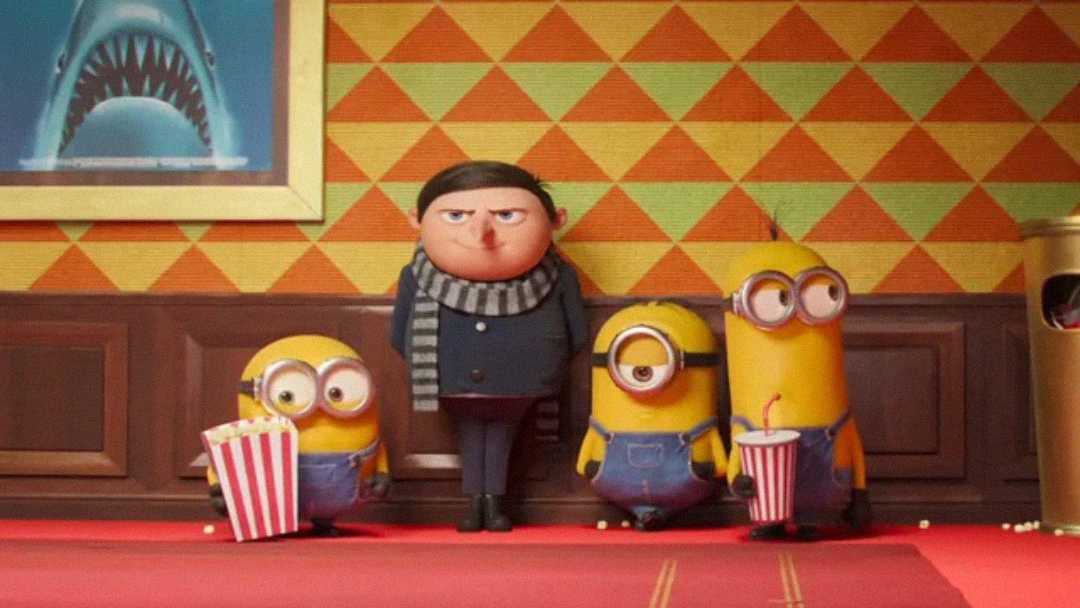 A serious-looking boy in black with a striped scarf stands against a wall next to three yellow Minions, two holding popcorn and soda, in a room with colorful wallpaper and a framed shark poster.