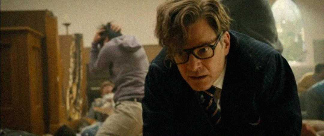 A man in a suit and glasses leans forward with a serious expression in a chaotic indoor setting, while people in the background appear to be in distress and taking cover.