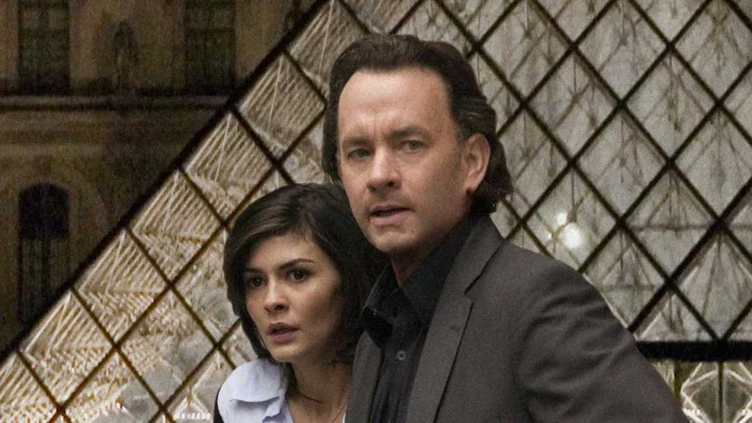 A man in a dark suit and a woman with short dark hair stand close together, looking concerned, in front of the glass pyramid entrance of the Louvre Museum.