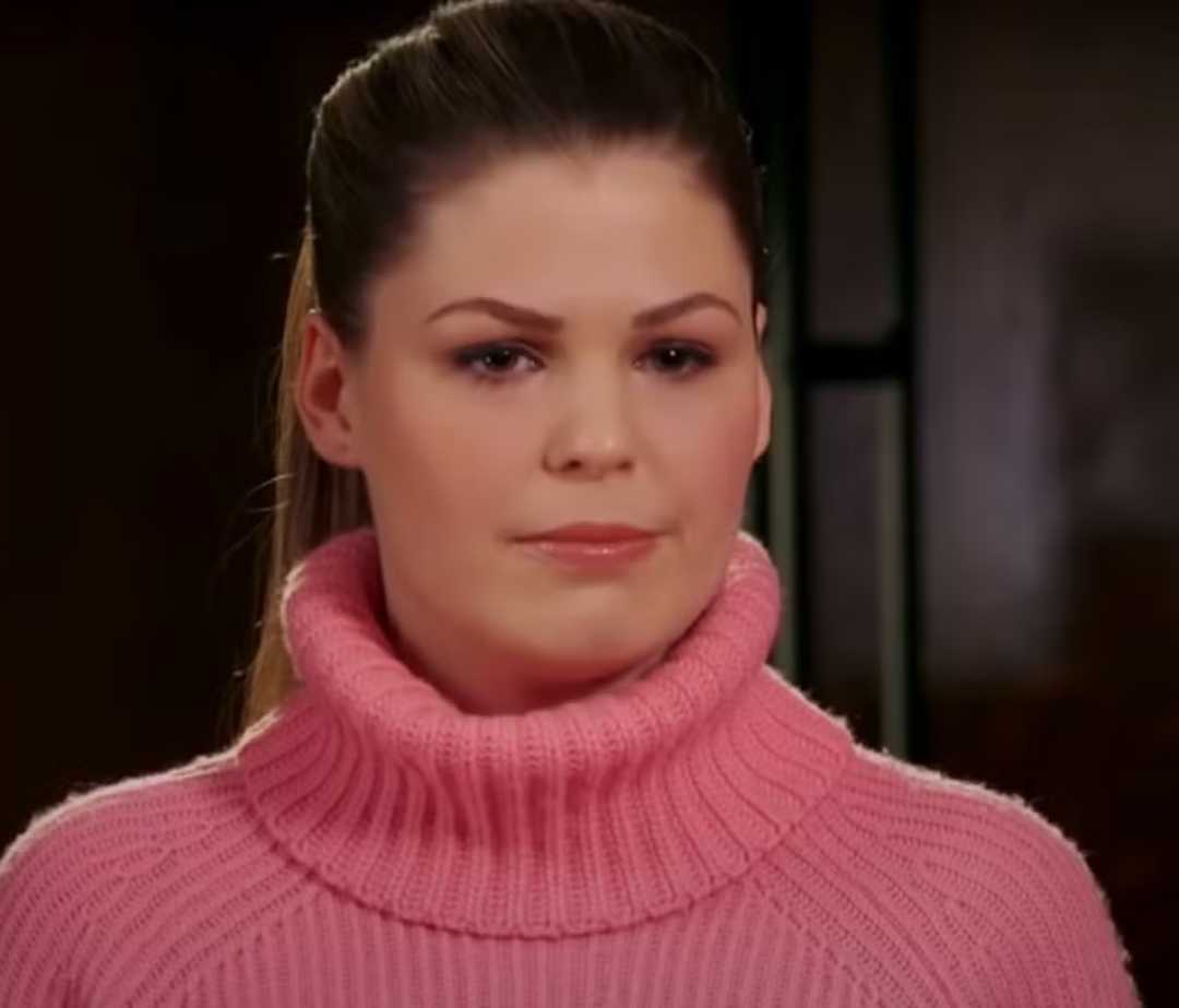 A woman with long brown hair tied back is wearing a thick pink turtleneck sweater, looking forward with a neutral expression. The background is softly blurred.