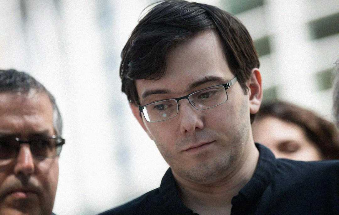 A man with short dark hair, wearing glasses and a black shirt, looks downward with a serious expression. There are blurred faces of other people beside him in the background.