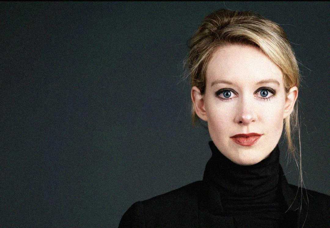 A woman with blonde hair tied back, wearing a black turtleneck and black blazer, looks directly at the camera against a dark, plain background.