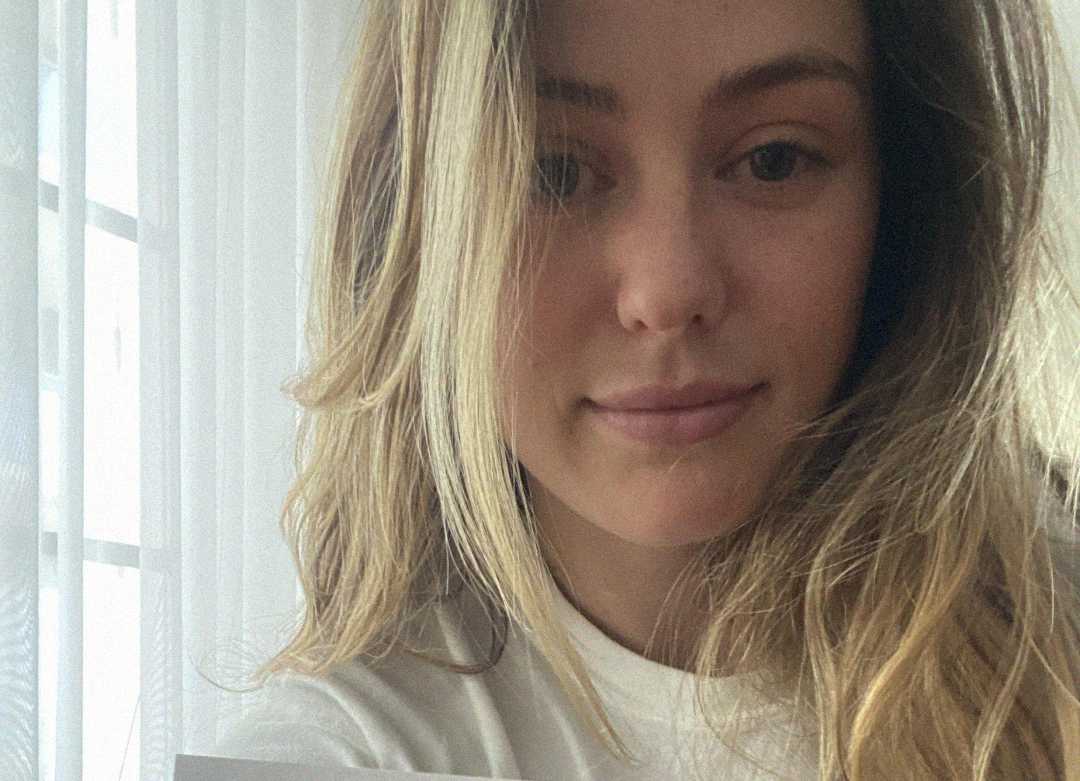 A young woman with long, wavy blonde hair takes a selfie indoors, standing near sheer white curtains with natural light coming through. She is wearing a white shirt and has a neutral expression.