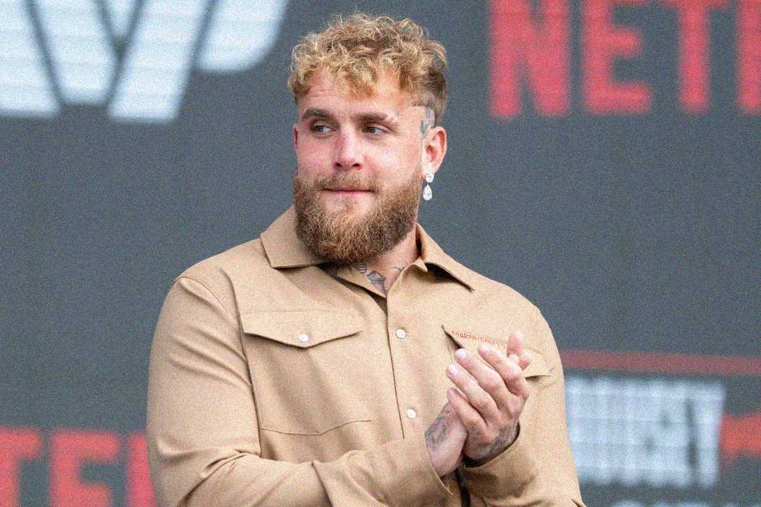 A man with curly blonde hair and a beard, wearing a tan button-up shirt, stands in front of a backdrop with blurred red and white text, clapping his hands and looking to the side.