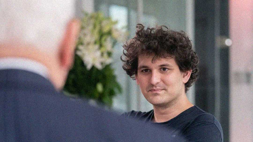 A young man with curly hair and a black shirt looks forward, while another person with gray hair is seen from behind. There is a blurred plant and bright indoor background.