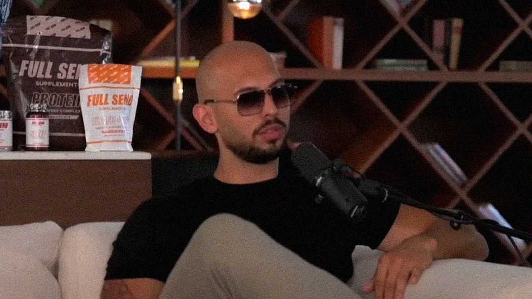 A bald man wearing sunglasses and a black shirt sits on a white couch, speaking into a microphone. Shelves with books and supplement bags are in the background.