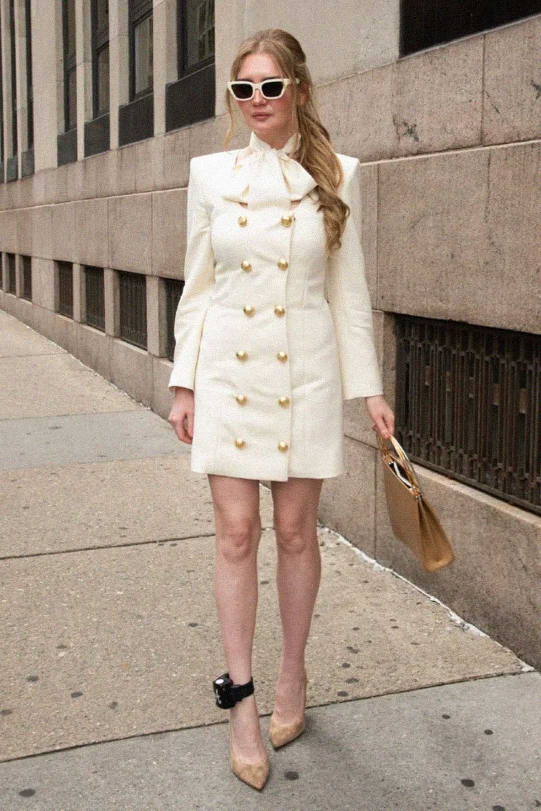 A woman stands on a city sidewalk wearing a white double-breasted coat dress with gold buttons, beige heels, white sunglasses, and holding a tan handbag. She has long, wavy hair and wears a black ankle monitor.
