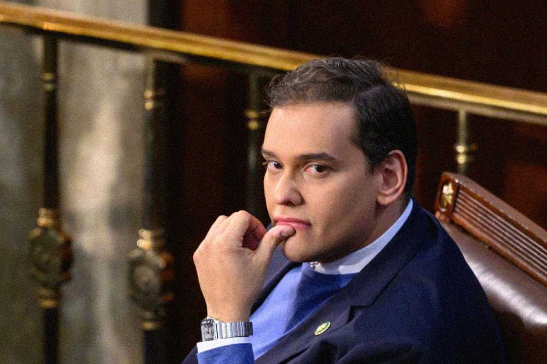 A person in a suit sits in a formal setting, resting their chin on their hand and looking thoughtfully toward the camera. There is a railing and blurred background behind them.