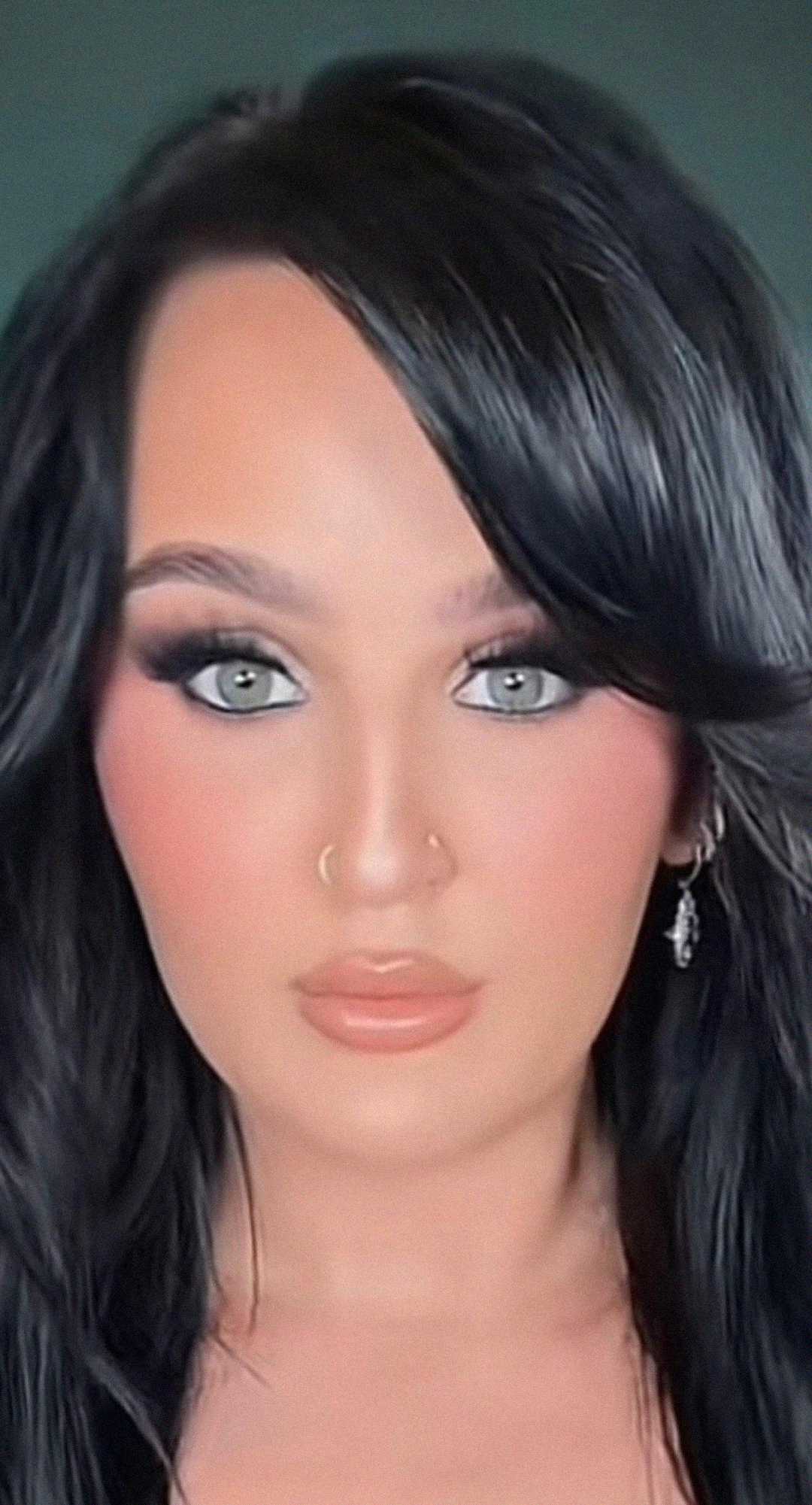 A person with long dark hair, light eyes, and flawless makeup poses against a dark background. They have a nose ring, a small earring, and a neutral facial expression.
