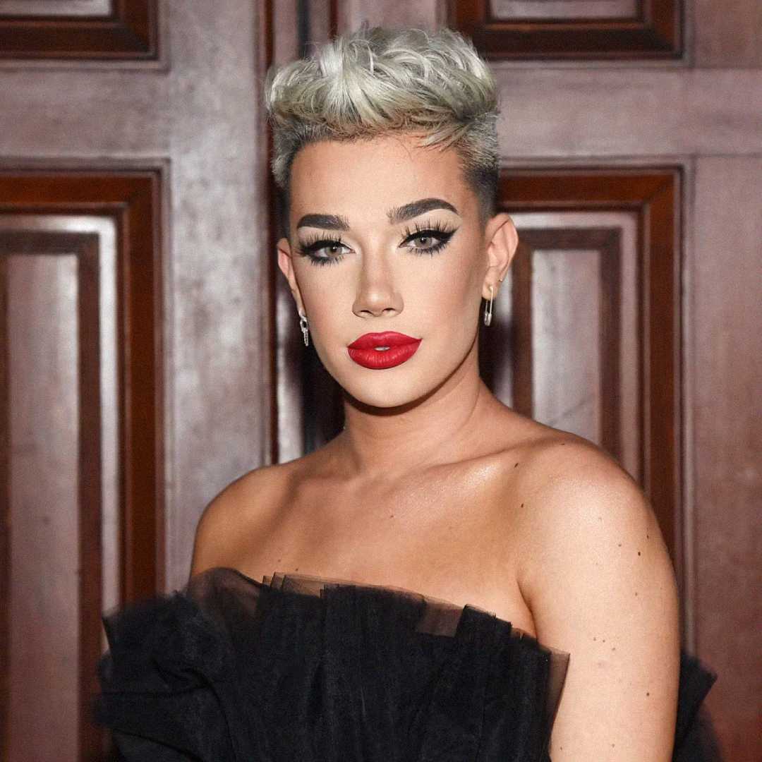 A person with short, styled platinum blonde hair, dramatic eye makeup, bold red lipstick, and wearing an off-the-shoulder black tulle outfit, poses in front of wooden panel doors.