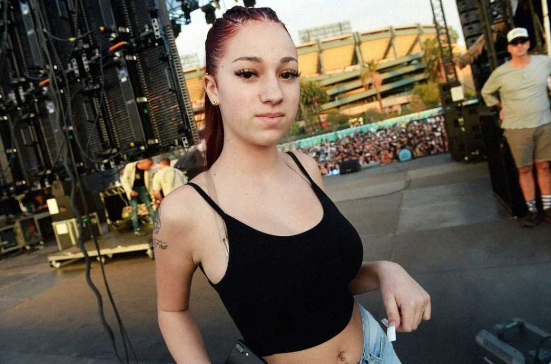 A young woman with long red hair in a black tank top stands on stage, holding a phone, with a crowd and stadium seating visible in the background during an outdoor event.