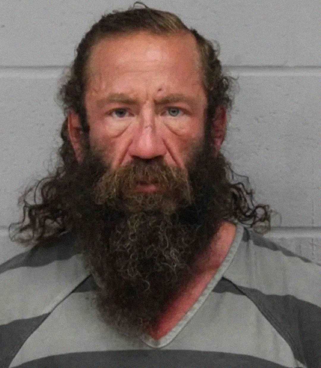 A man with a long, curly brown beard and wavy hair looks directly at the camera, wearing a gray-striped shirt, standing against a light gray cinder block wall.
