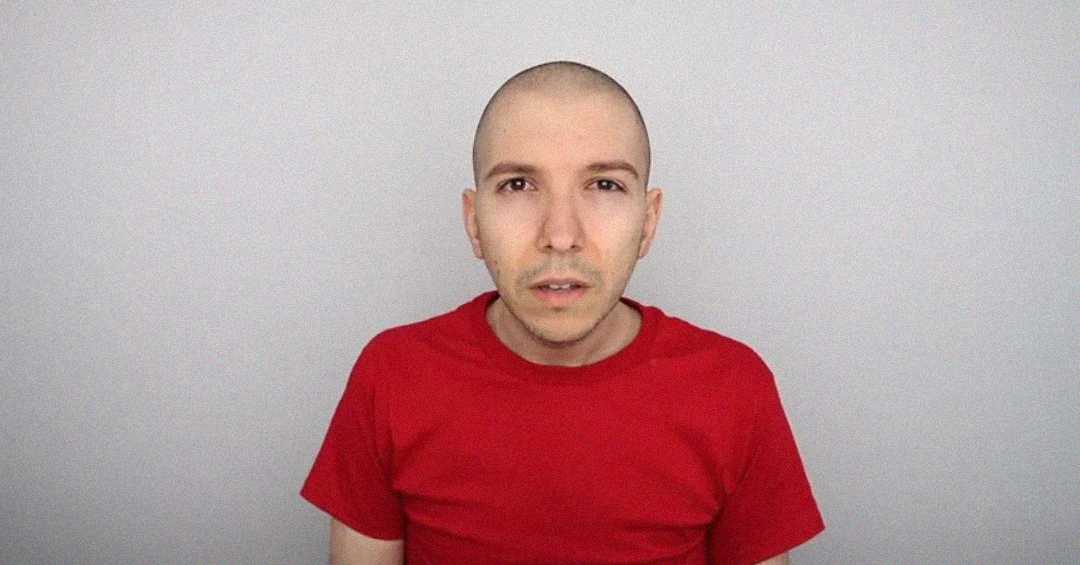 A person with a shaved head wearing a plain red t-shirt stands against a light gray background, looking directly at the camera with a neutral expression.