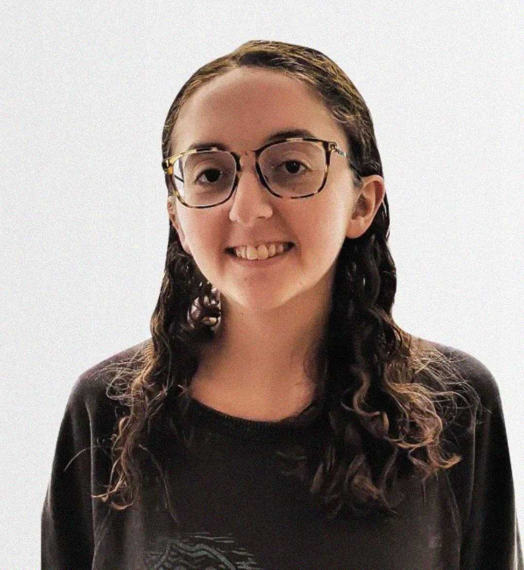 A person with long, curly brown hair and glasses is smiling while wearing a dark shirt. The background is plain and light-colored.