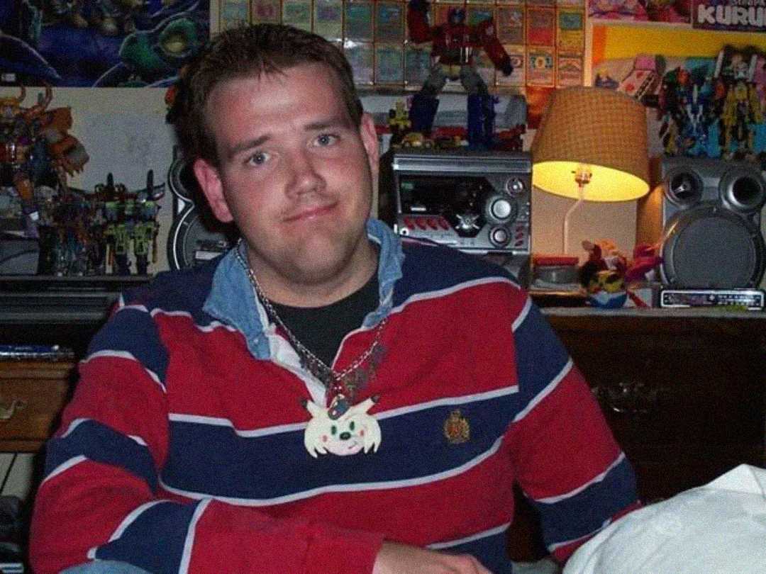A young man in a red and blue striped shirt sits in a cluttered room filled with toys and collectibles, wearing a necklace with a white cartoon character pendant and looking at the camera with a slight smile.