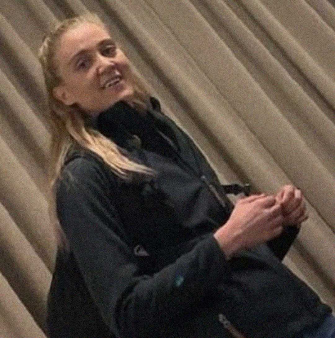 A person with long blonde hair, wearing a dark jacket and backpack, stands in front of beige curtains, looking slightly toward the camera with a subtle smile.