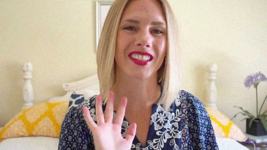 A woman with straight blonde hair and red lipstick smiles and waves at the camera. She is wearing a blue patterned blouse and is seated in a bright room with yellow pillows and a plant in the background.